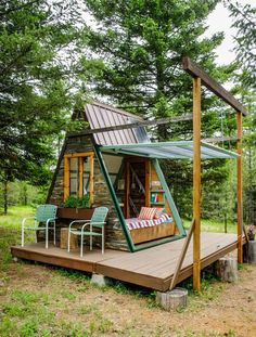 a small cabin in the woods with a bed and chairs on the deck, surrounded by trees