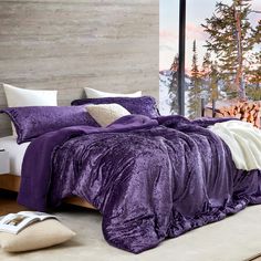 a bed with purple comforter and pillows in front of a window overlooking the mountains