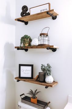 two wooden shelves with plants and pictures on them in a white bathroom, next to a toilet