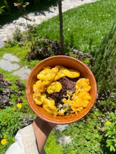 a person holding up a bowl filled with bananas and other things in the grass next to some flowers