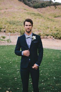 a man in a tuxedo poses for a photo on the grass with his hands in his pockets