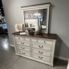 a white dresser with drawers and a mirror on the top, in front of a gray wall