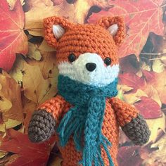 a crocheted stuffed animal with a scarf around it's neck sitting in front of autumn leaves