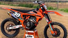 an orange dirt bike parked on top of a metal stand in the dirt next to a fence