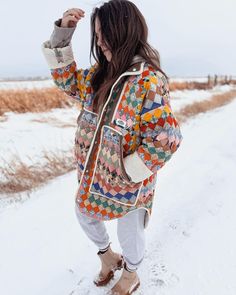 a woman standing in the snow with her hand up to her ear and wearing a multicolored jacket