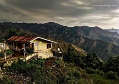 a small house on the side of a hill with mountains in the background and cloudy skies