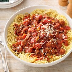 a bowl of spaghetti with meat sauce and parmesan cheese on top, next to silverware