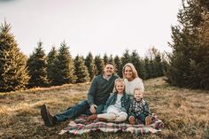 a family sitting on a blanket in the middle of a field surrounded by pine trees