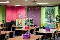 a classroom filled with lots of desks covered in purple and green bins next to windows