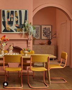 a dining room with pink walls and yellow chairs around a wooden table in front of a fireplace