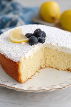 a slice of lemon cake on a plate with blueberries and lemons in the background