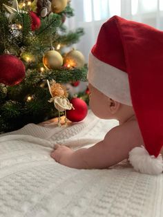 a baby wearing a santa hat sitting in front of a christmas tree with ornaments on it