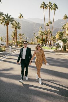 a man and woman walking down the street holding hands with palm trees in the background