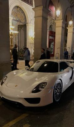 a white sports car parked in front of a tall building with people standing around it