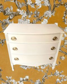 a white dresser sitting in front of a yellow wall with flowers on the wall behind it