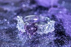 a heart shaped diamond ring sitting on top of a purple surface with water droplets around it