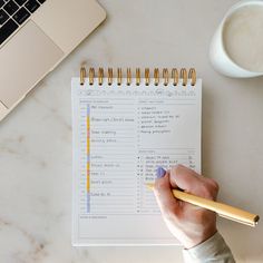 a person holding a pencil next to a notebook on top of a white table with a laptop