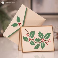 two cards with holly designs on them sitting on a table next to some pine cones