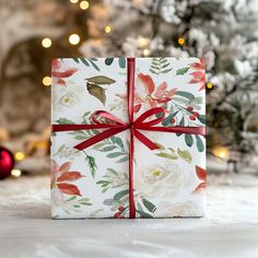 a gift wrapped in white paper with red ribbon and flowers on it, next to a christmas tree