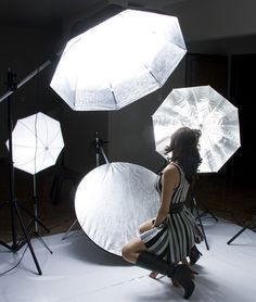 a woman is sitting in front of several umbrellas on a white surface with lights behind her