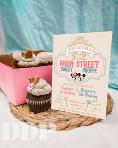 two cupcakes and a sign on a wicker basket for a sweet shoppe