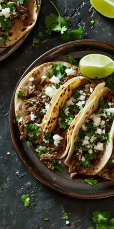 three tacos on a plate with limes and cilantro next to them