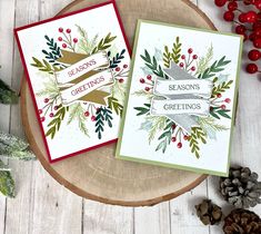 two christmas cards sitting on top of a wooden table next to pine cones and berries