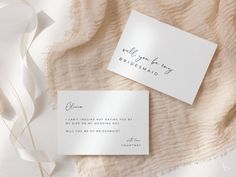 two white wedding cards with calligraphy on them sitting next to a pile of fabric