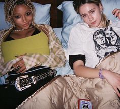 two young women laying in bed with one holding a purse and the other looking at the camera