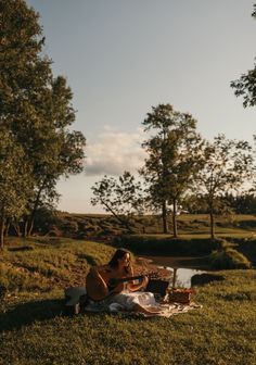 a woman sitting on top of a lush green field next to a river playing an acoustic guitar