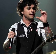 a man holding a microphone while standing in front of a guitar and wearing a vest