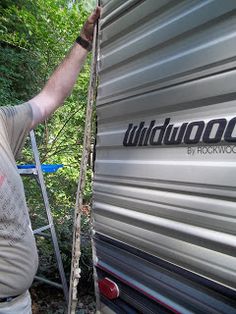 a man standing next to a silver trailer in the woods with his hand on the ladder