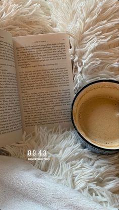 an open book sitting on top of a white blanket next to a cup of coffee