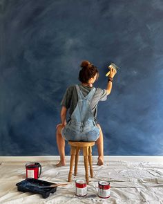 a woman sitting on top of a stool holding a paint roller in front of her face