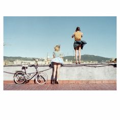 two women are sitting on a wall and one is standing next to a bike while the other looks at her