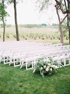 an outdoor ceremony set up with white chairs and flowers on the grass in front of trees