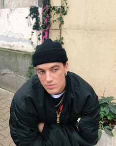 a young man sitting on the ground wearing a black jacket and beanie, with his hands in his pockets