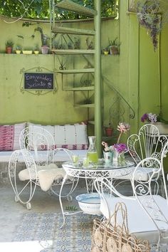 an outdoor patio with white furniture and flowers on the table, next to a green wall