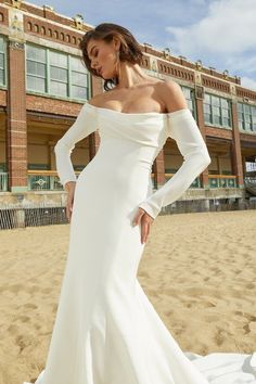 a woman in a white wedding dress standing on the beach with her hands on her hips