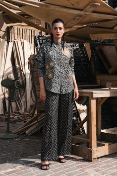 a woman standing in front of a wooden structure wearing black and white patterned pants with gold accents