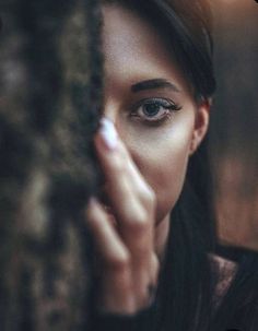 a woman with blue eyes is looking out from behind a tree trunk and holding her hand to her face