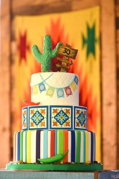 a multi - tiered cake with colorful decorations and cactus on top is displayed in front of a brightly colored background