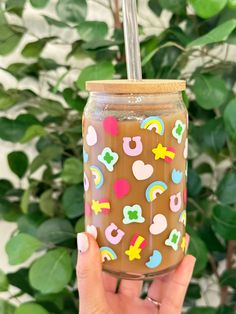 a hand holding up a mason jar with rainbows and hearts on it, in front of a green plant