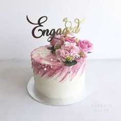 a white cake with pink flowers and the word engaged on top is decorated with gold lettering