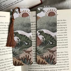 two bookmarks with dinosaurs on them are sitting next to an open book and one has a tassel