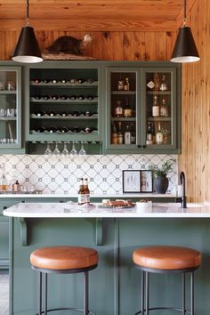 a kitchen with green cabinets and stools next to a bar area that has wine glasses on it