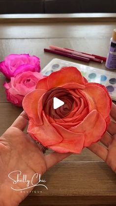 a person holding a flower in their hand with paint and watercolors on the table