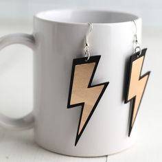 a pair of wooden earrings with black and gold lightning bolt design on them, hanging from a white mug