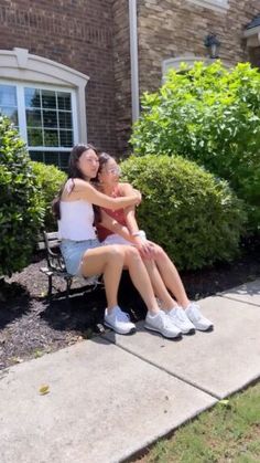 two young women sitting on a bench in front of a brick house, one holding her arm around the other's neck