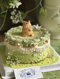 there is a green cake with a bear decoration on the top and flowers in the background